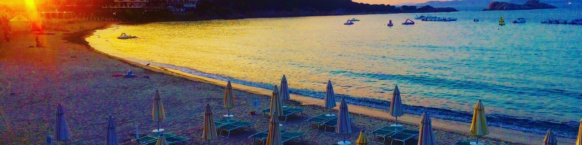 A beach with rows of sun loungers and umbrellas, a calm sea, and a sunset in the background.