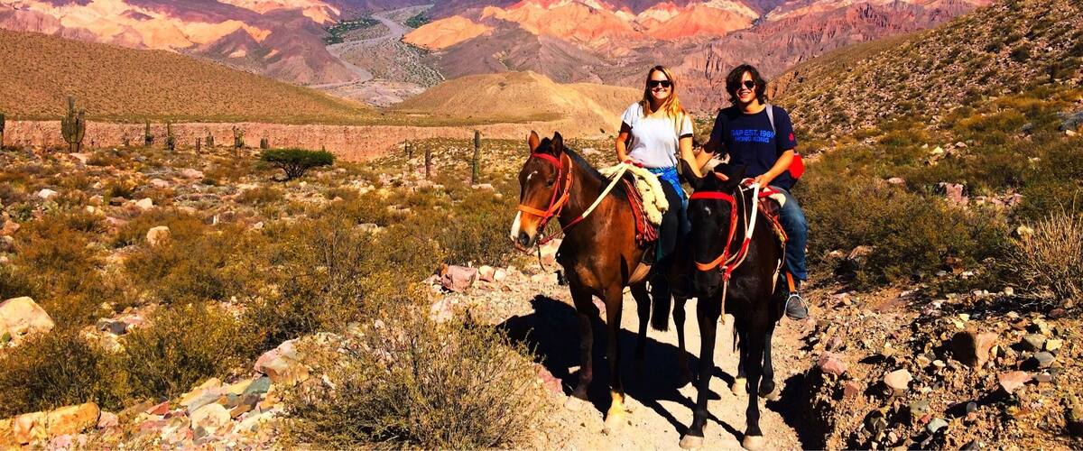 Two people riding horses in a desert landscape with mountains in the background.