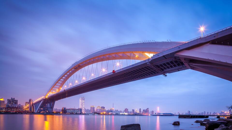 Eine große Brücke überspannt einen ruhigen Fluss, dessen Oberfläche die Lichter der Stadt reflektiert.