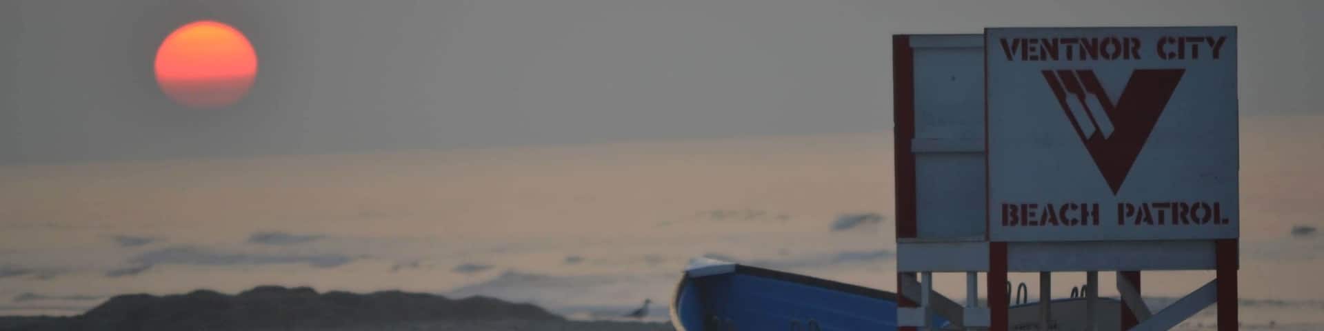 A beach patrol sign and a boat on the beach with the sun setting in the background.