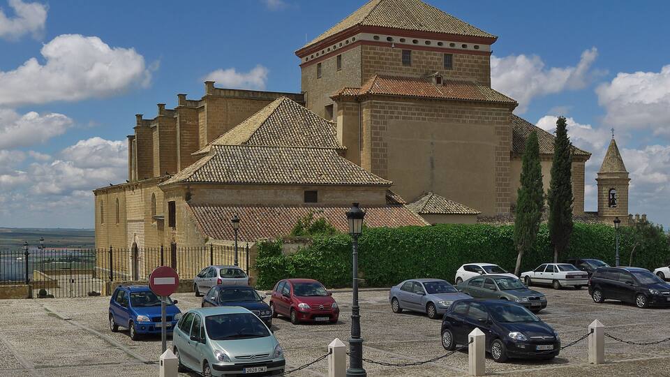 A historic building with a tiled roof and a tower, surrounded by parked cars and a clear sky.