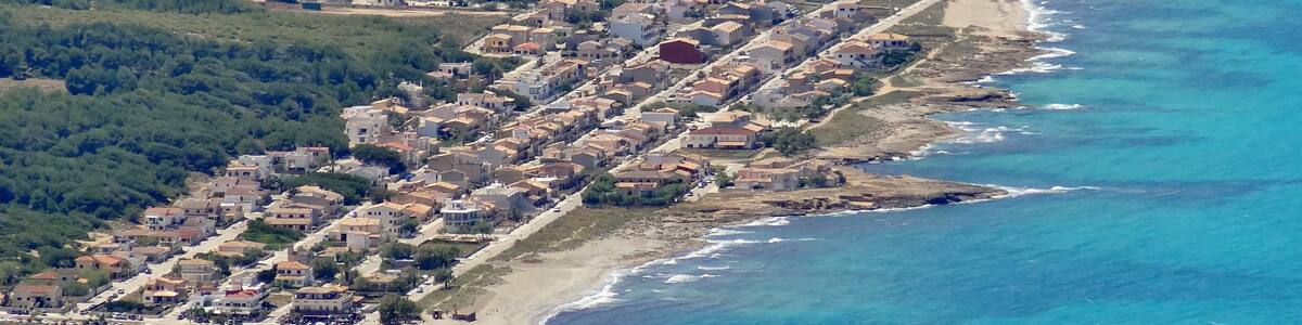 An aerial view of a coastal town with dense housing along the shoreline, a sandy beach, and a backdrop of dense forest.