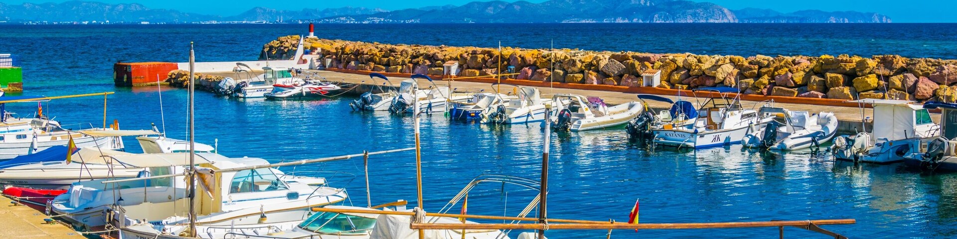 A marina with various boats docked, a rocky breakwater, and a clear blue sky.