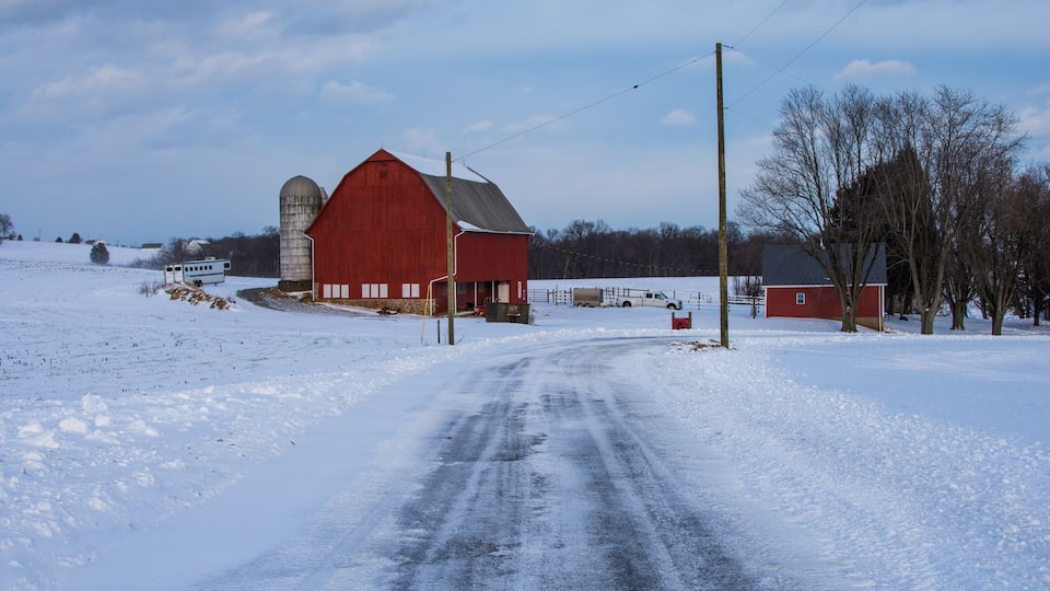 A snow-covered rural road leading to a red barn with a silo.