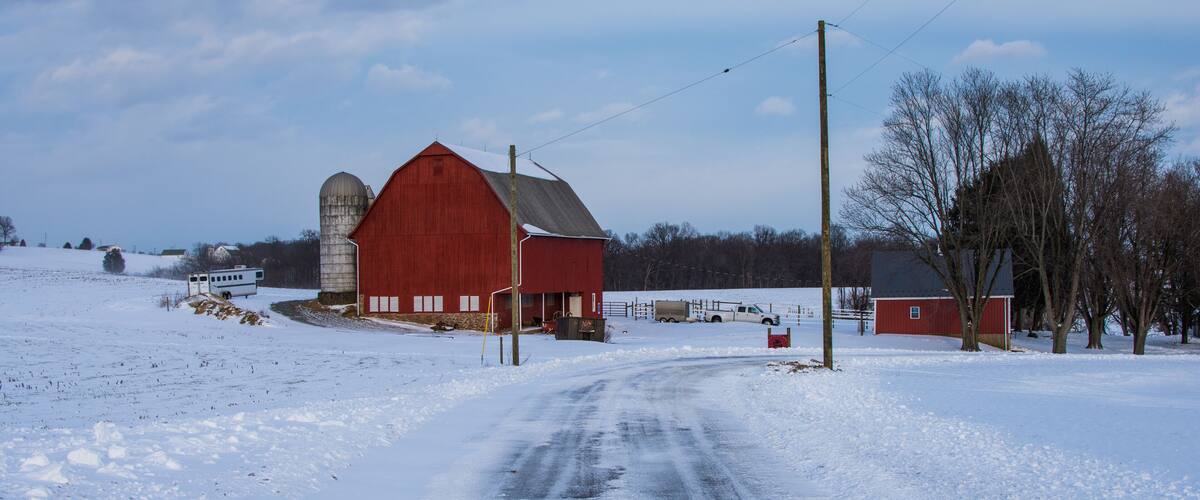 A snow-covered rural road leading to a red barn with a silo.