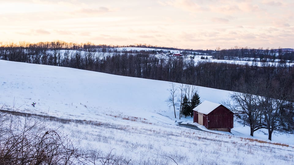 A snow-covered landscape with a red barn, trees, and a distant village.