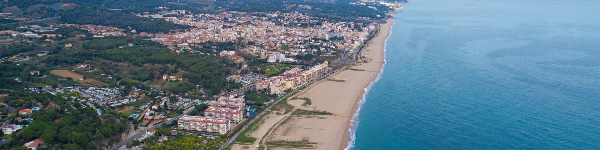Vista aérea de uma cidade costeira com uma extensa praia, área urbana densa e colinas ao redor.