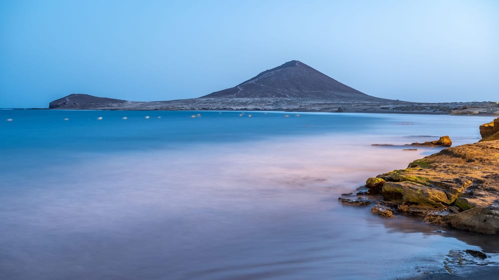 A coastal landscape with a prominent mountain, calm sea, and rocky shoreline.