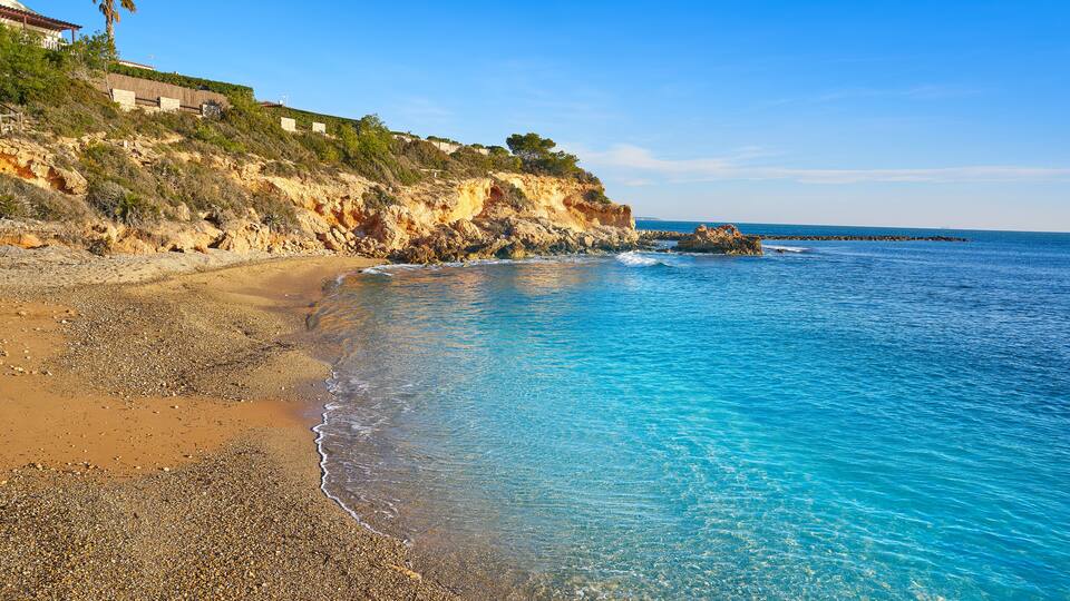 A coastal landscape with a pebbly beach, clear blue waters, and a rocky cliff with vegetation.