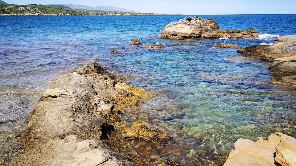A rocky shoreline with clear blue water and distant hills.