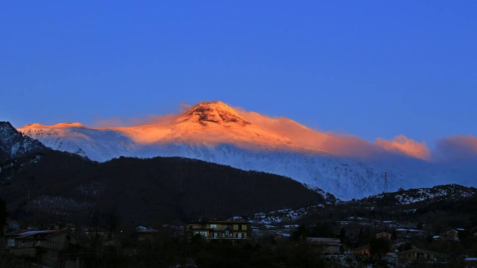 A snow-capped mountain at sunset with a village in the foreground.