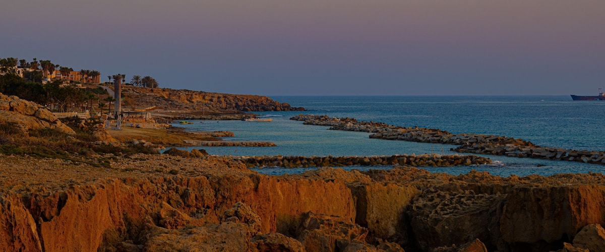A rocky coastline with a ship in the distance and buildings on the left.