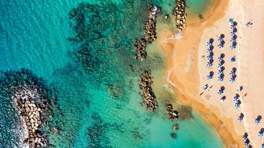 An aerial view of a beach with clear turquoise waters, rocky formations, and sun umbrellas.