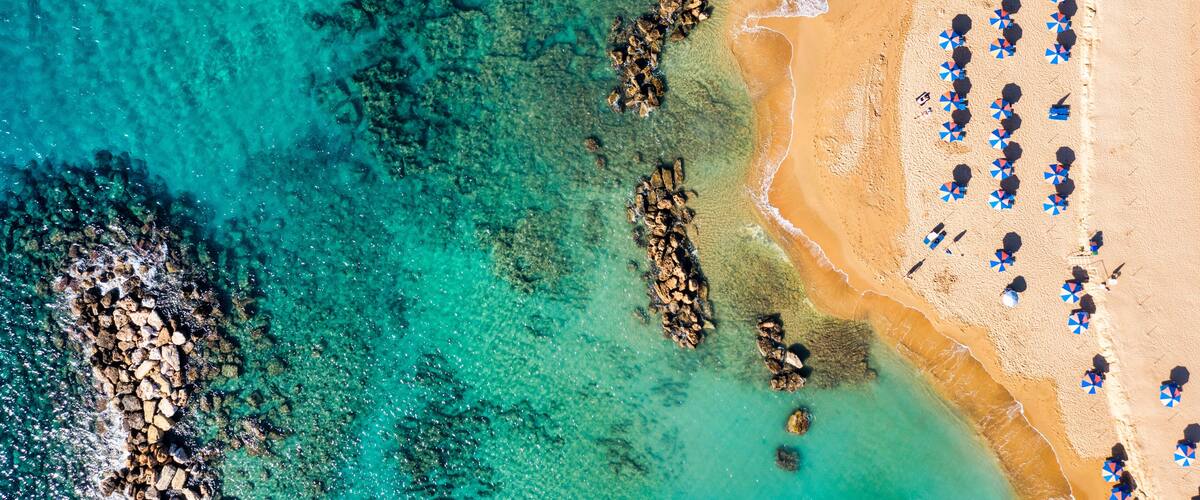 An aerial view of a beach with clear turquoise waters, rocky formations, and sun umbrellas.