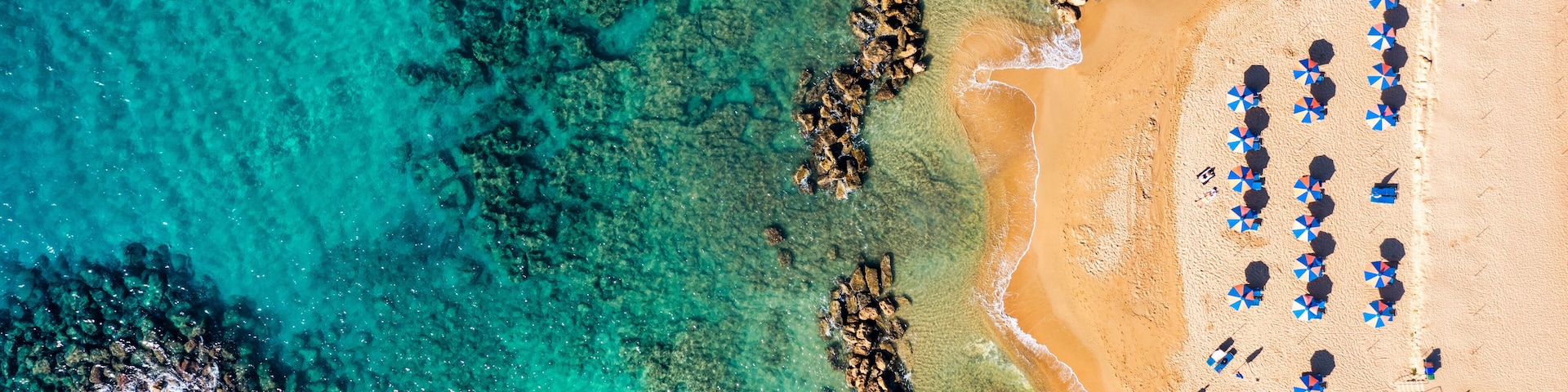 An aerial view of a beach with clear turquoise waters, rocky formations, and sun umbrellas.