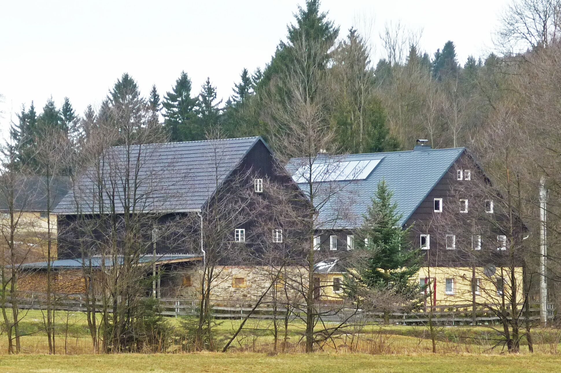 A large stone house with a steep roof and multiple chimneys, surrounded by trees and a fence.