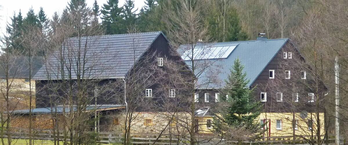A large stone house with a steep roof and multiple chimneys, surrounded by trees and a fence.