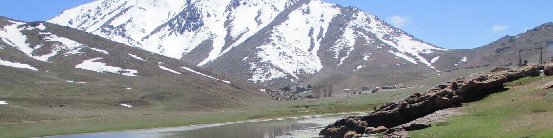 A mountain with snow-capped peaks, a clear lake reflecting the sky, and a rocky shoreline.