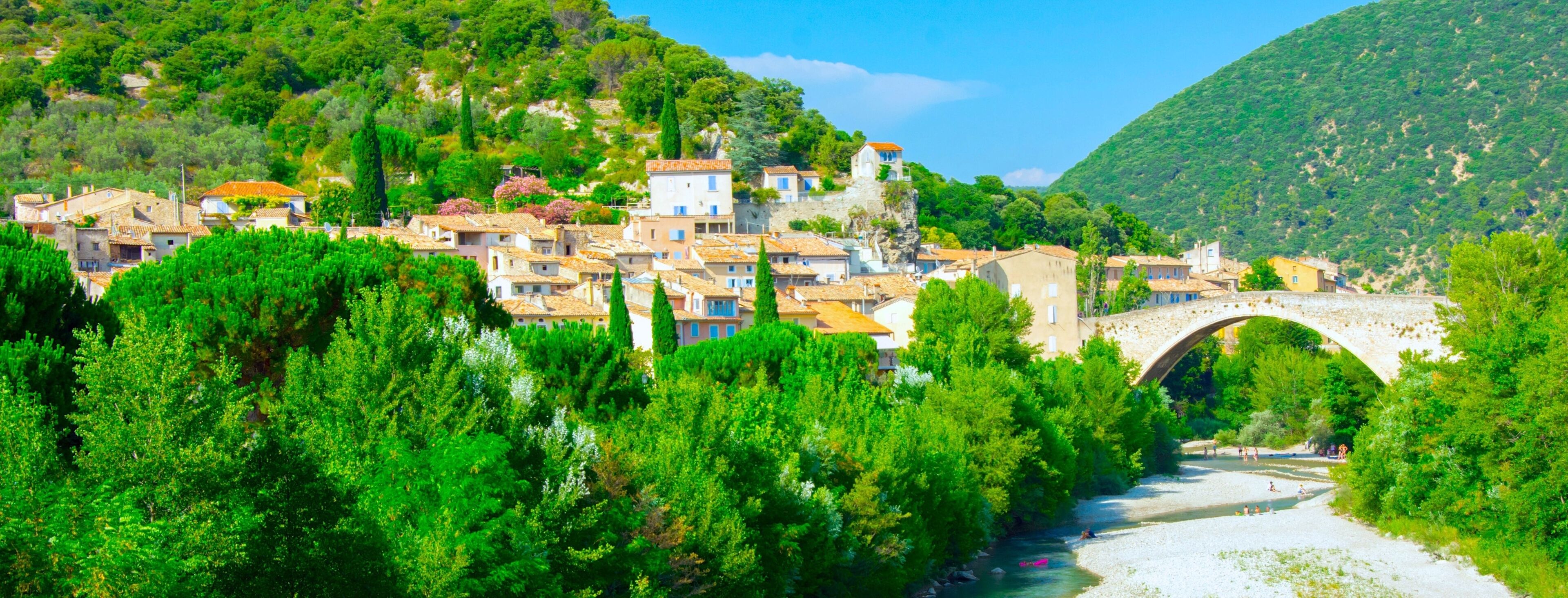A village with a stone bridge over a river, surrounded by lush green trees and hills.