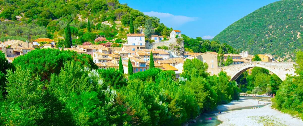 A village with a stone bridge over a river, surrounded by lush green trees and hills.