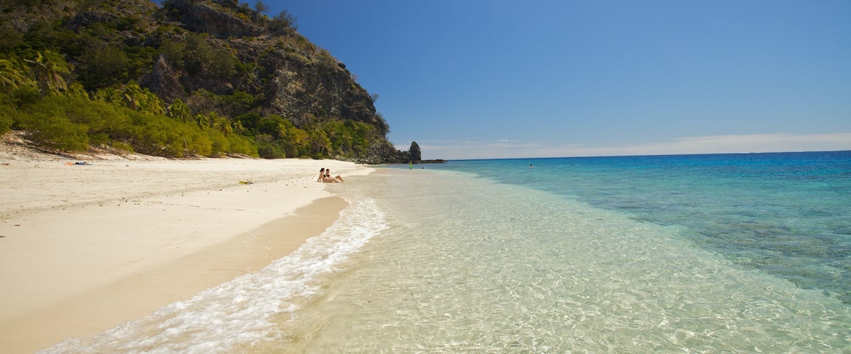 A beach with clear turquoise water, sandy shore, and a rocky cliff with vegetation.