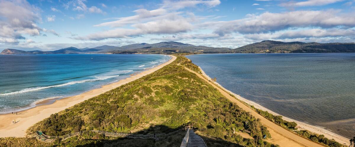 A coastal landscape with a sandy beach, a grassy path, and a calm sea under a partly cloudy sky.
