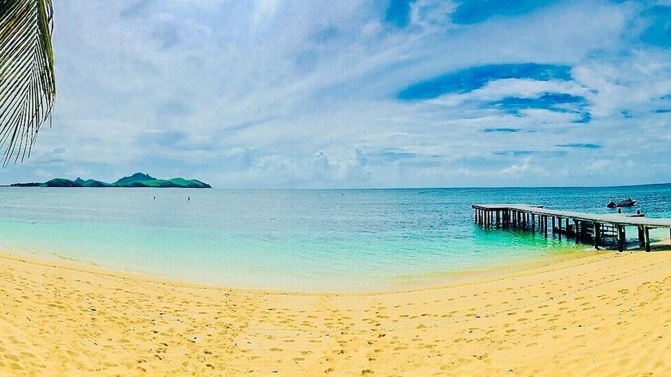 A beach with a wooden pier extending into calm turquoise waters, a clear sky, and distant islands.
