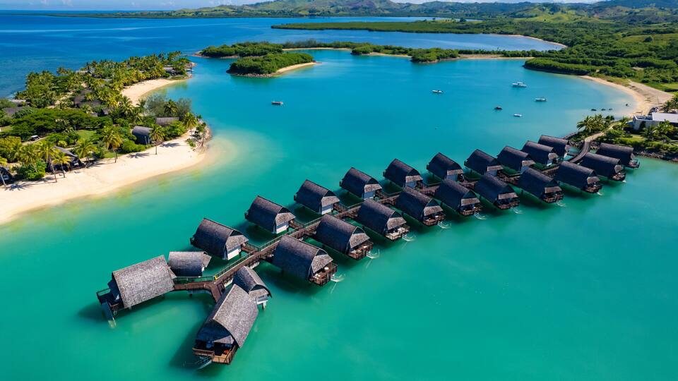 An aerial view of overwater bungalows in turquoise waters, surrounded by lush greenery and a sandy beach.