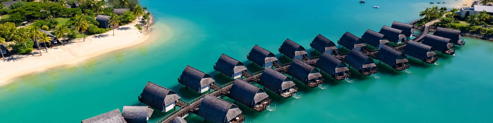 Vista aérea de bungalows sobre el agua en aguas turquesas, rodeados de vegetación exuberante y una playa de arena.