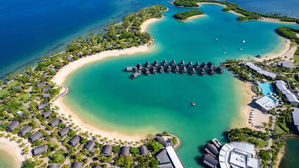An aerial view of a resort with overwater bungalows, a beach, and clear turquoise waters.
