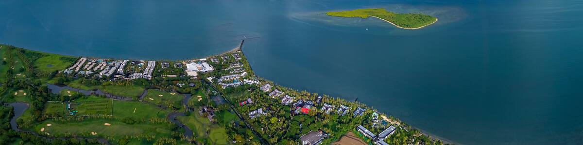 Expansive aerial view of a luxurious tropical resort and golf course on the coast, with a small, verdant island just offshore under a vast, cloud-filled sky