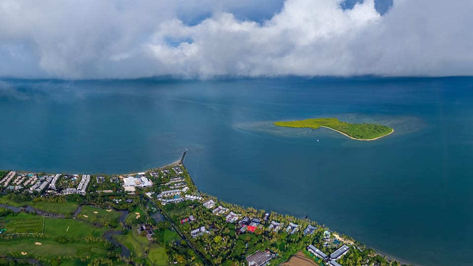 Expansive aerial view of a luxurious tropical resort and golf course on the coast, with a small, verdant island just offshore under a vast, cloud-filled sky