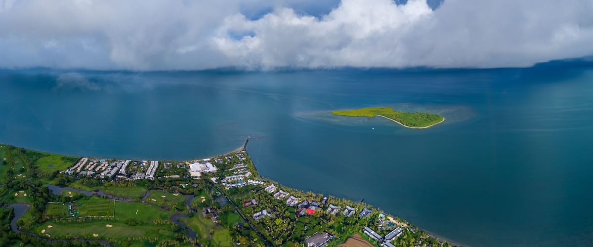 Expansive aerial view of a luxurious tropical resort and golf course on the coast, with a small, verdant island just offshore under a vast, cloud-filled sky