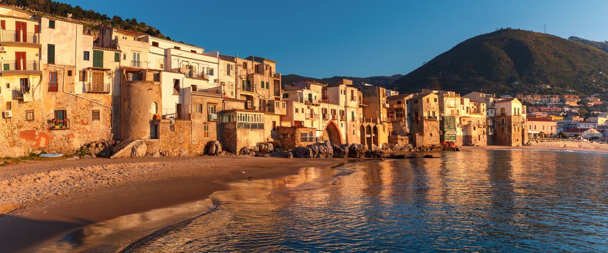 A coastal town with buildings along the shore, a clear sky, and a hill in the background.
