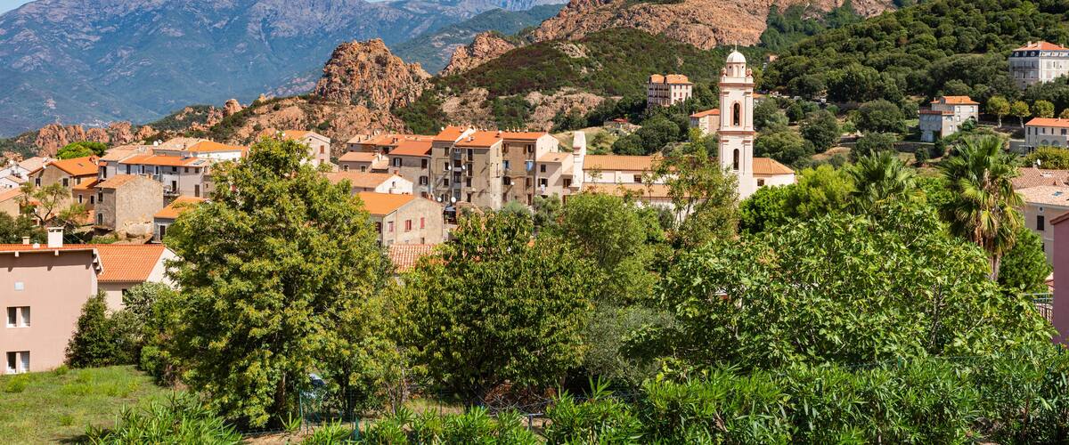 A town with red-roofed buildings, a prominent church tower, and mountains in the background.