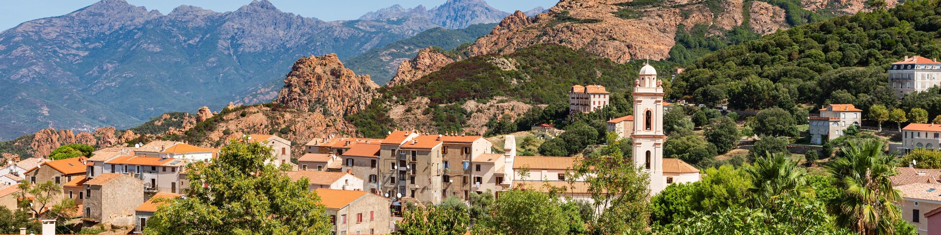 A town with red-roofed buildings, a prominent church tower, and mountains in the background.