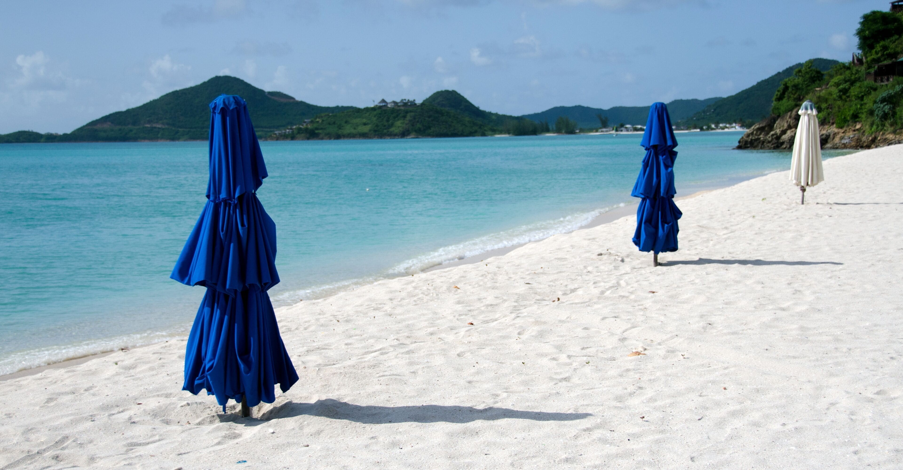 A beach with two blue umbrellas and a white umbrella on the sand, with a clear blue sea and green hills in the background.