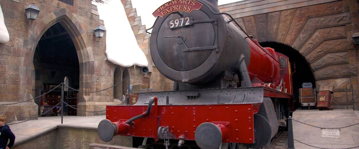Platform view of a red steam train in front of a themed entrance at Universal Studios Florida during a sunny day