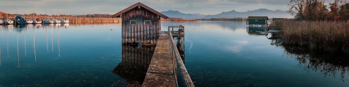 A wooden boathouse on a dock extending into a calm lake, surrounded by mountains and autumn foliage.