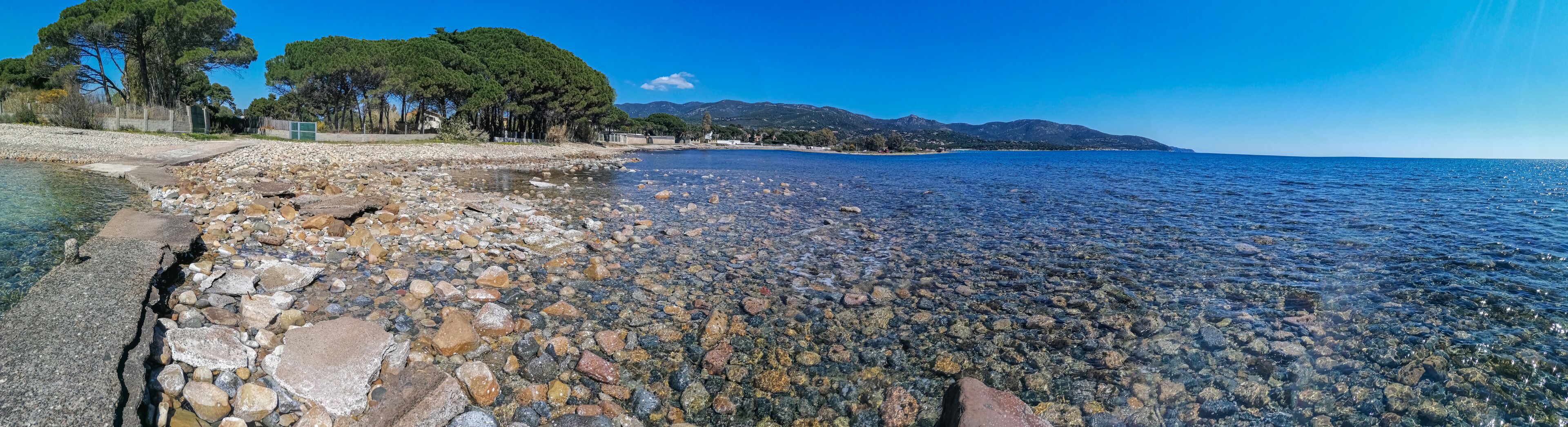 A rocky shoreline with clear water, trees, and distant mountains.