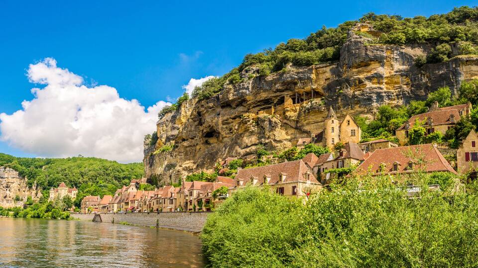 A river with a bridge, a town with red-roofed buildings, and a cliff with greenery.