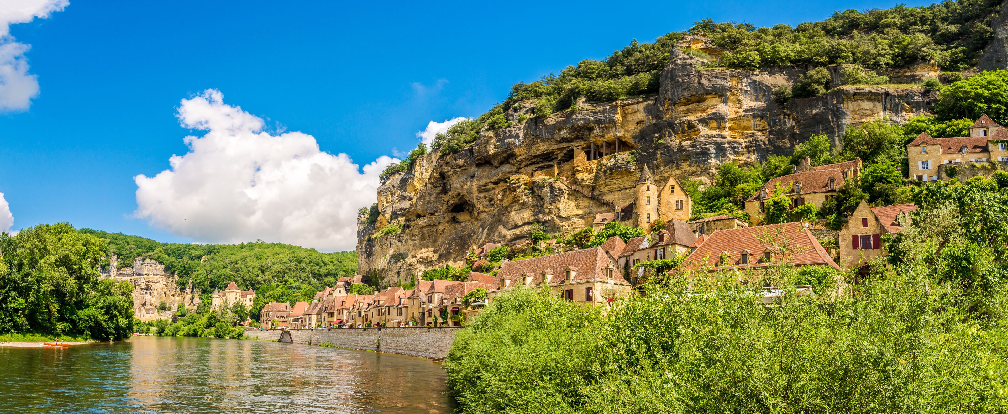 A river with a bridge, a town with red-roofed buildings, and a cliff with greenery.