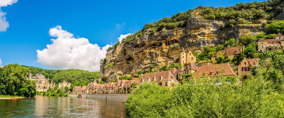 A river with a bridge, a town with red-roofed buildings, and a cliff with greenery.