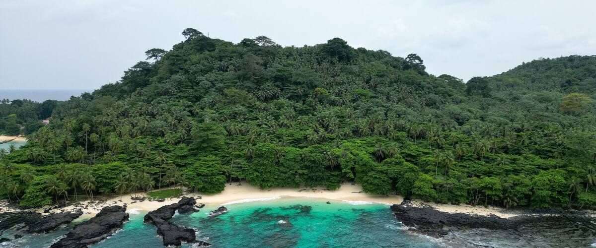 Uma praia tropical com águas turquesa cristalinas, cercada por floresta verde exuberante e afloramentos rochosos.