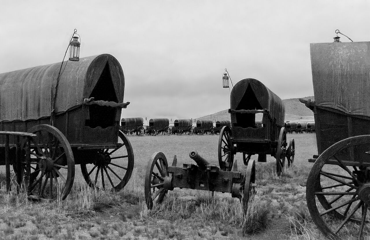 A row of covered wagons with lanterns, set in a field with a distant hill.
