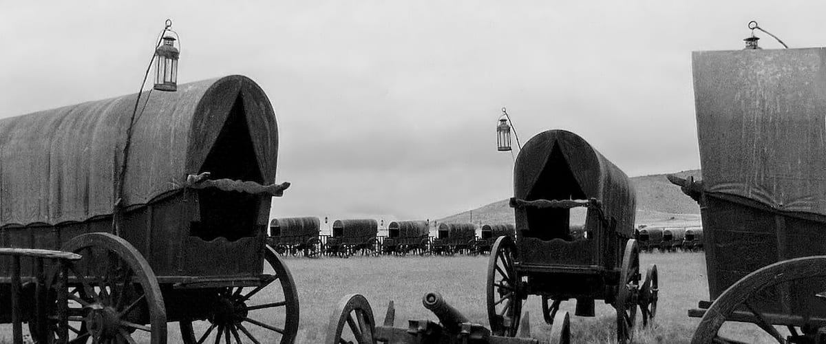 A row of covered wagons with lanterns, set in a field with a distant hill.