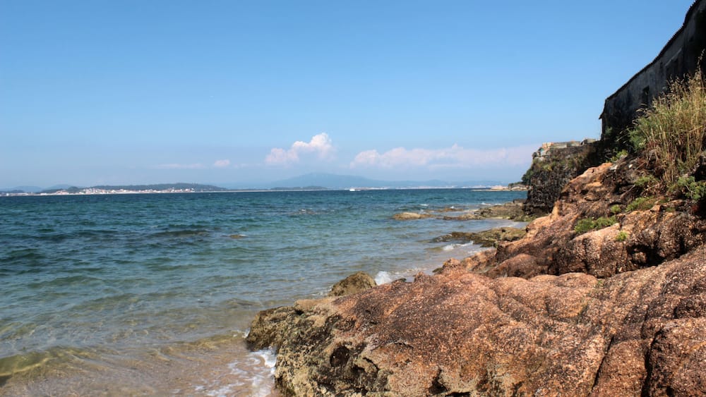 A rocky coastline with clear blue water and distant mountains.