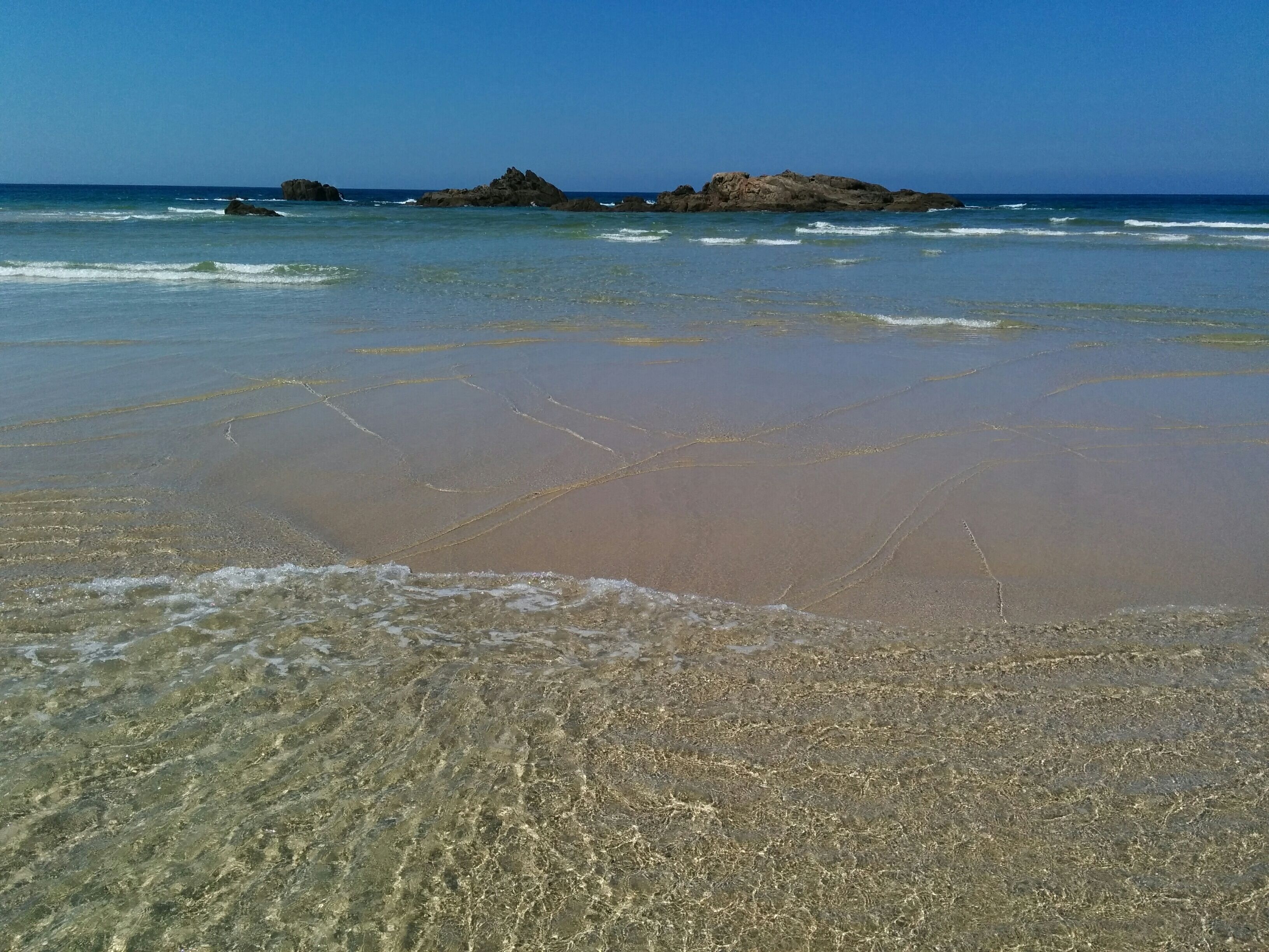 A beach with clear shallow waters, gentle waves, and rocky formations in the distance.