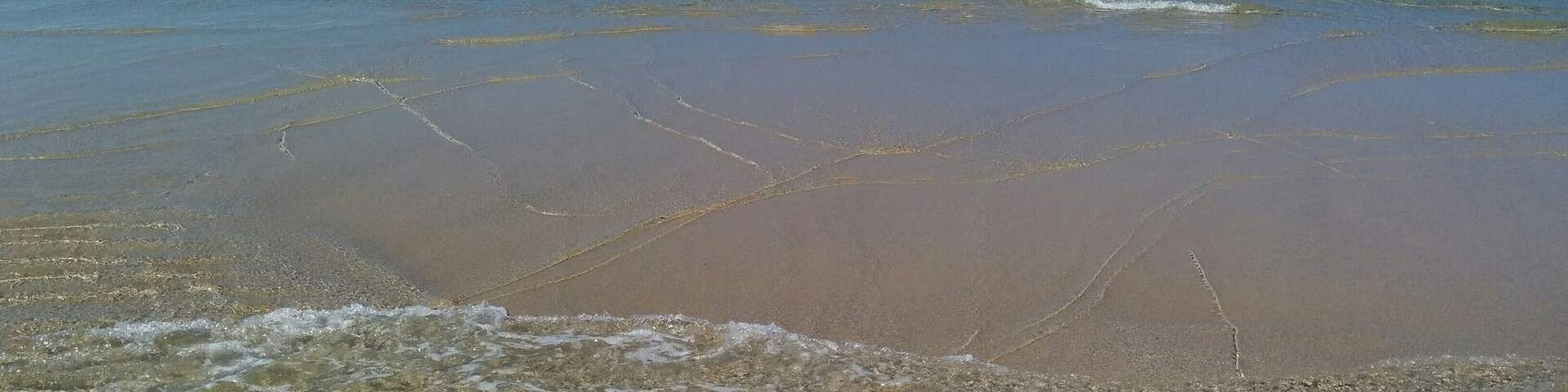 A beach with clear shallow waters, gentle waves, and rocky formations in the distance.