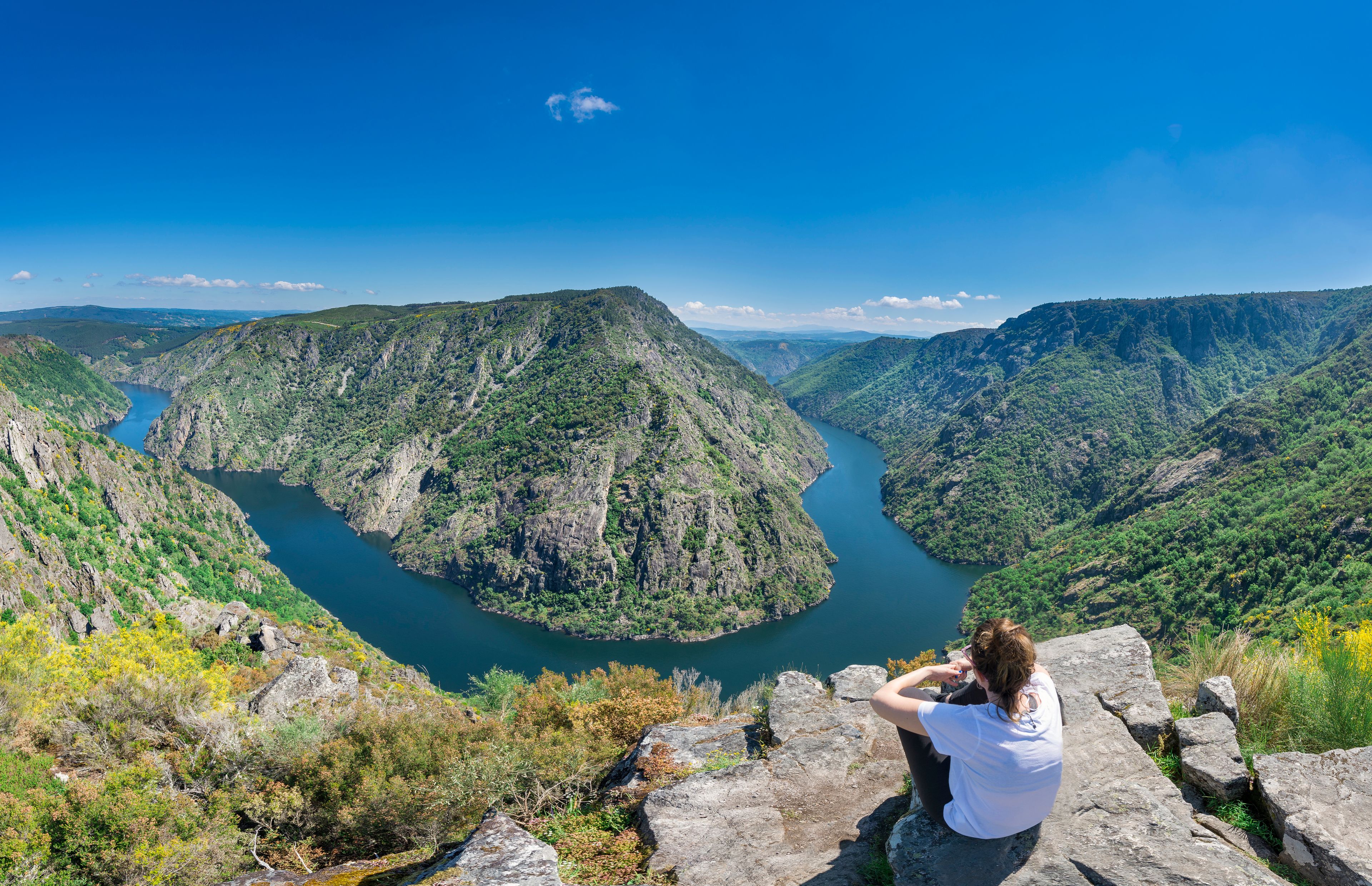 A person sitting on a rock overlooking a deep canyon with a river.